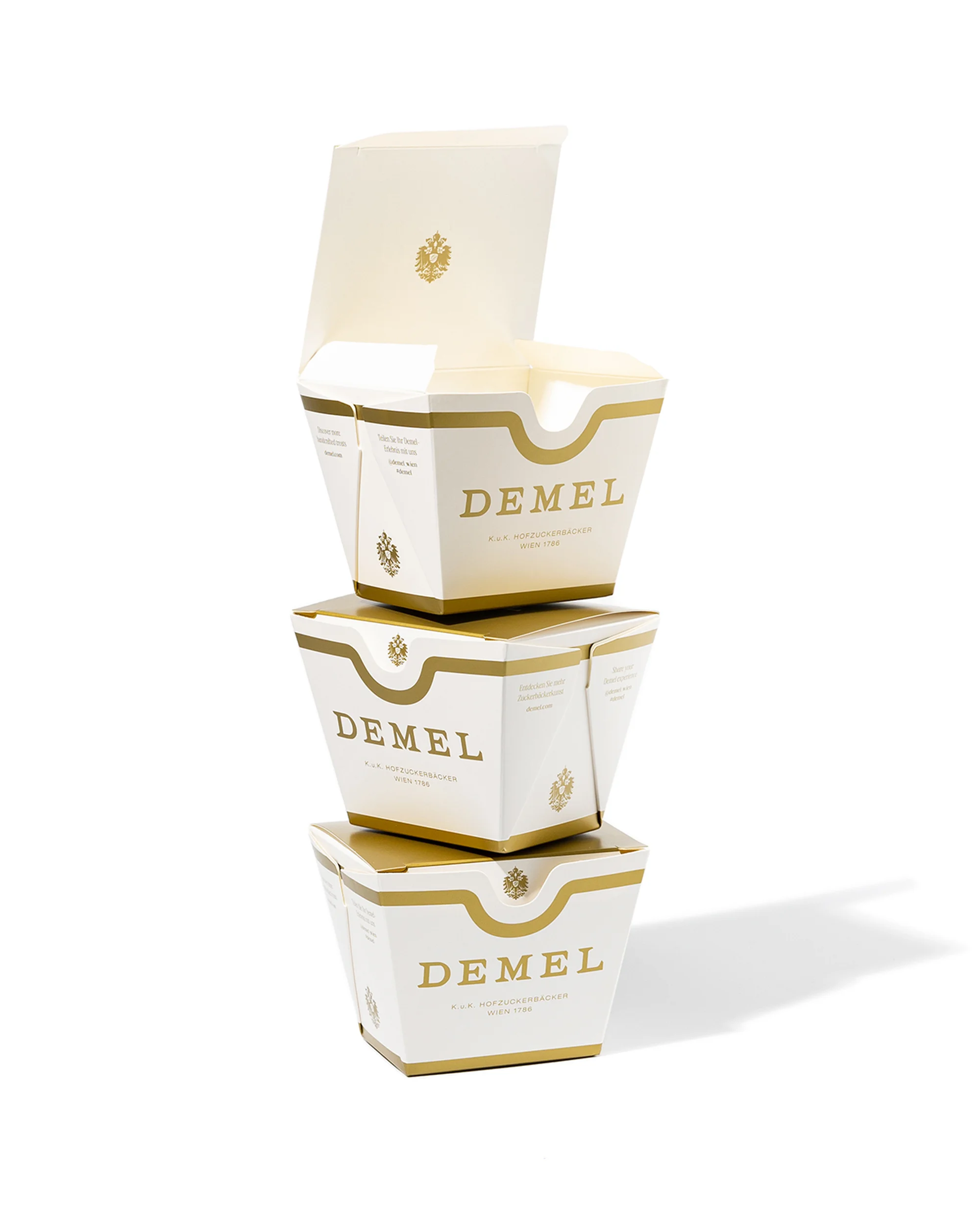 Three Demel boxes stacked on a white surface. The top box has its lid open, and the boxes have gold accents and logos.