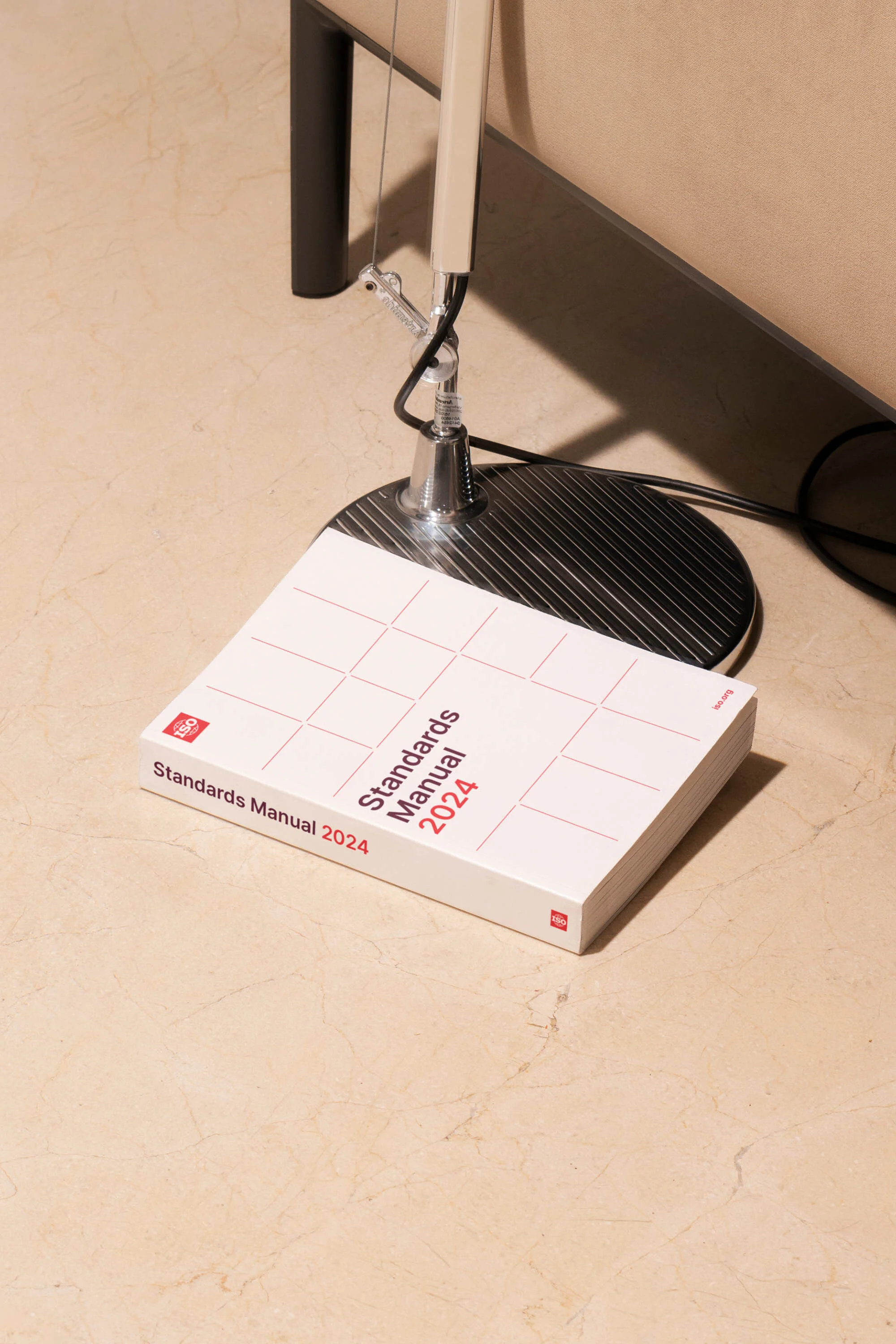 Product shot of a white 'Standards Manual 2024' book laying on a cream floor, lit by a modern adjustable chrome floor lamp.