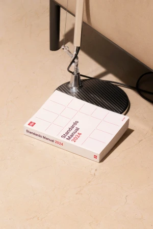 Product shot of a white 'Standards Manual 2024' book laying on a cream floor, lit by a modern adjustable chrome floor lamp.