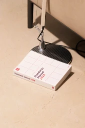 A white Standards Manual 2024 book placed on a beige floor beside a modern chrome desk lamp.
