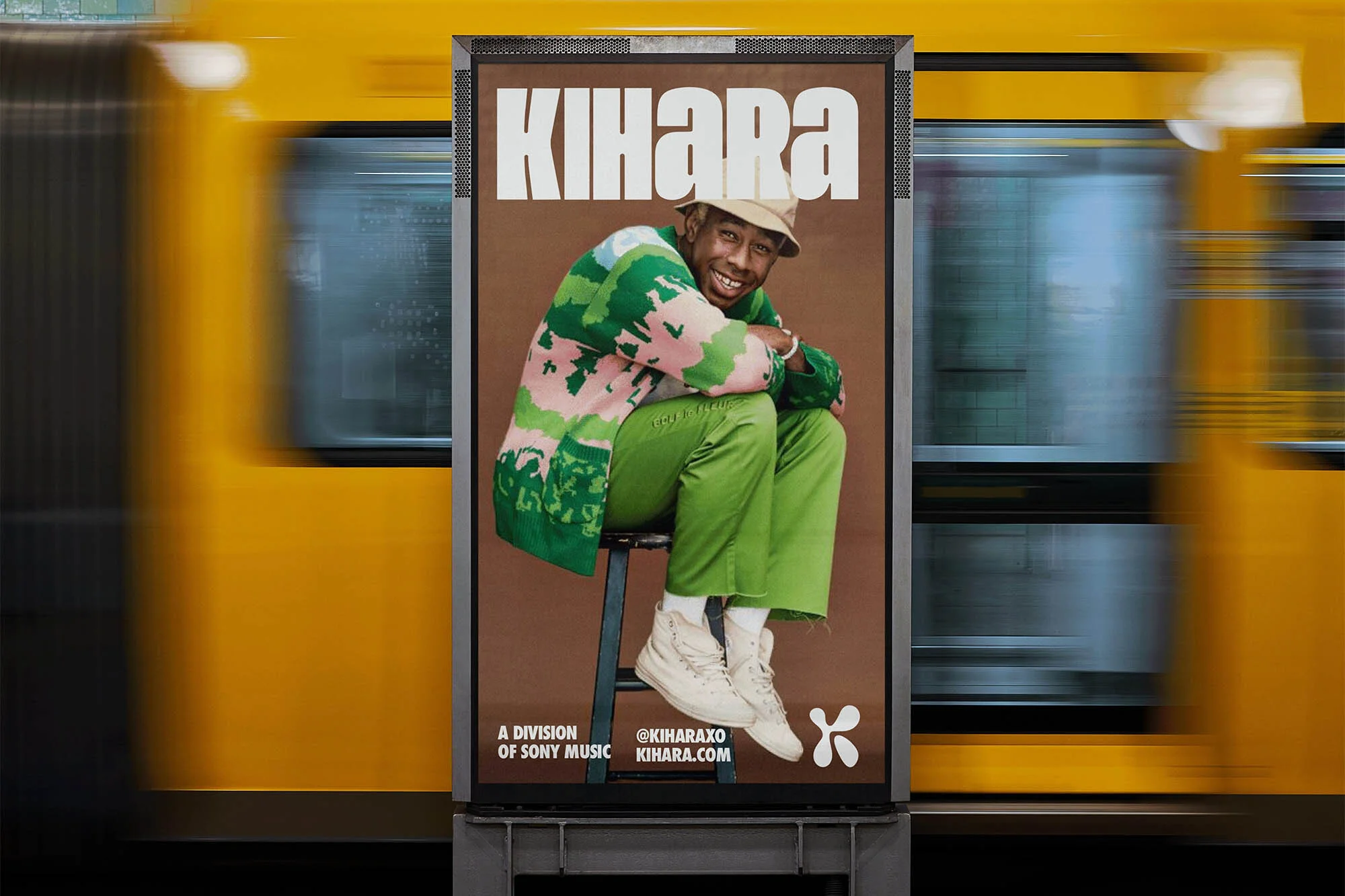 Poster featuring a person in green sweater and pants sitting on a stool, with the word KIHARA above them, next to a passing train.