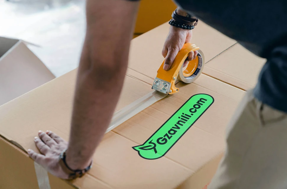 Adult sealing a large cardboard box with clear tape using a yellow dispenser. Box features a green logo on a plain indoor backdrop.