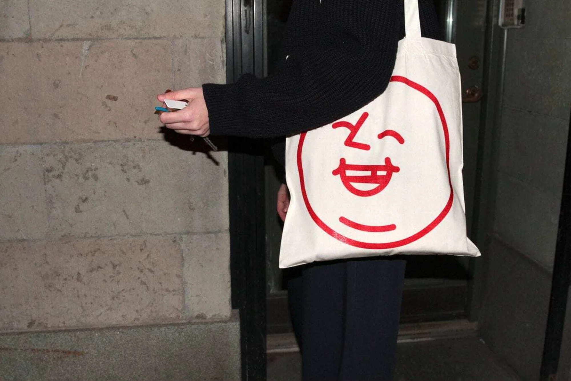Person holds keys near a door, a cream-colored tote bag with a red smiling face graphic is visible in a nighttime urban setting.