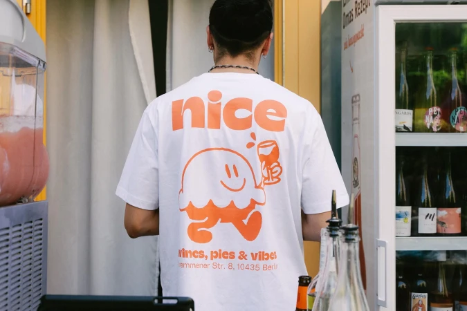 Man wearing a white t-shirt with an orange graphic stands near a drink dispenser and bar with wine bottles in the background.