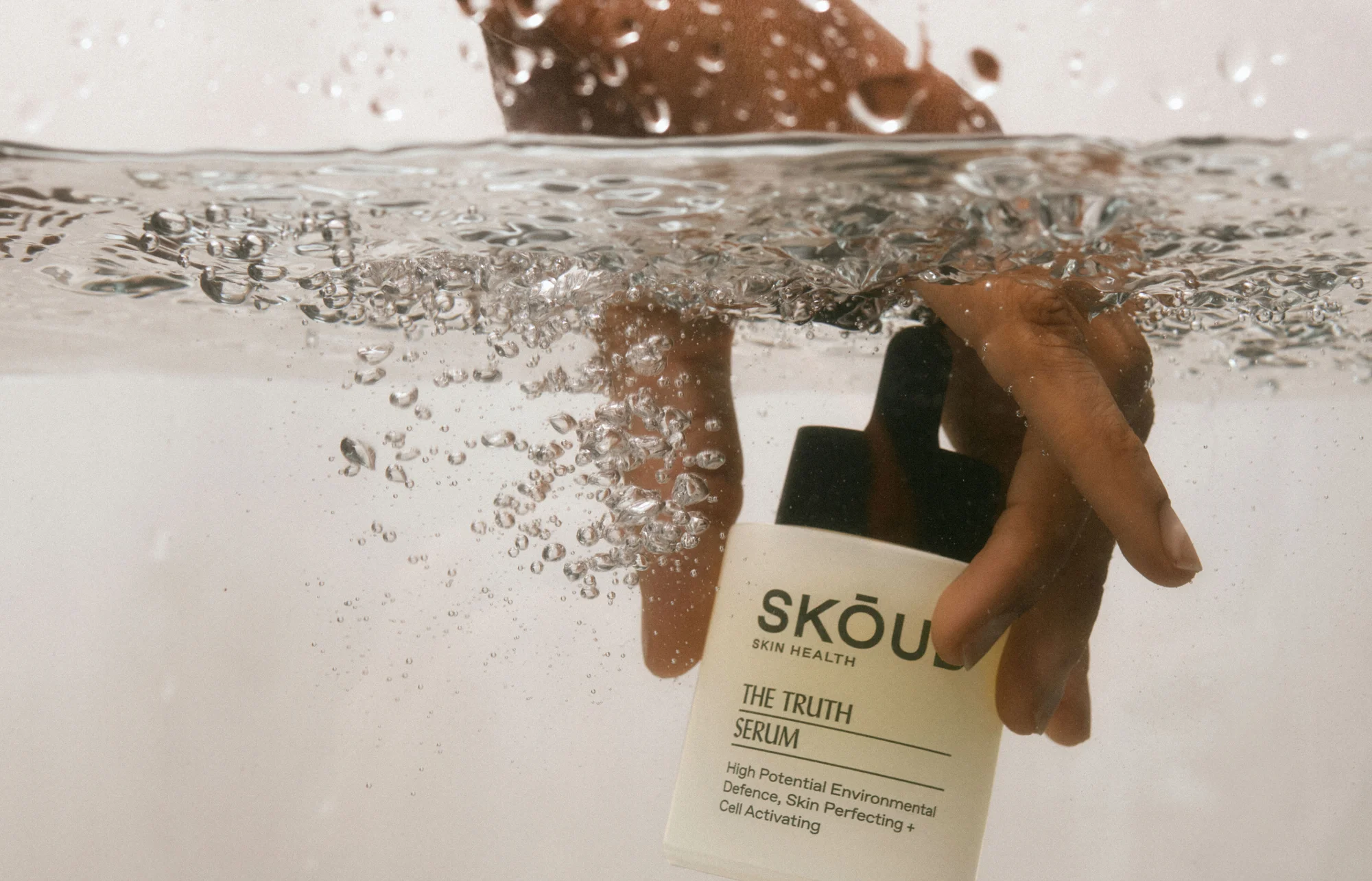Hand holding a Skou Skin Health serum bottle in water with bubbles, product shot, close-up of the label, skincare.
