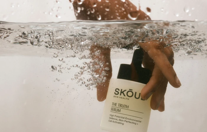 Hand holding a Skou Skin Health serum bottle in water with bubbles, product shot, close-up of the label, skincare.
