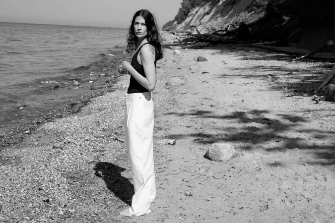 Model standing on a stony beach wearing a black tank top and white trousers by SKOUD Cosmetics