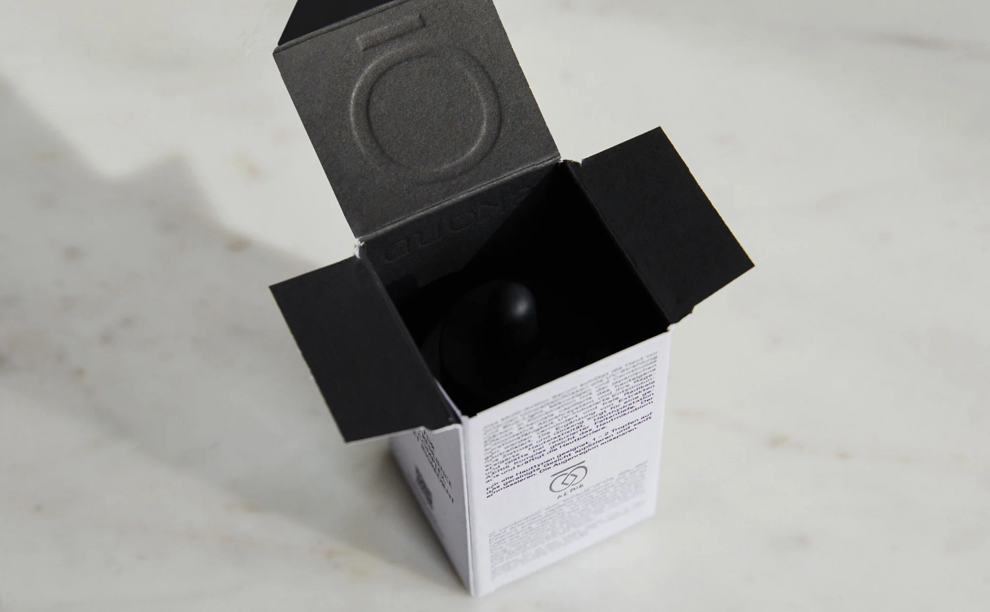Cosmetic product in a black and white box with the lid open on a neutral surface, revealing a dark dropper inside.