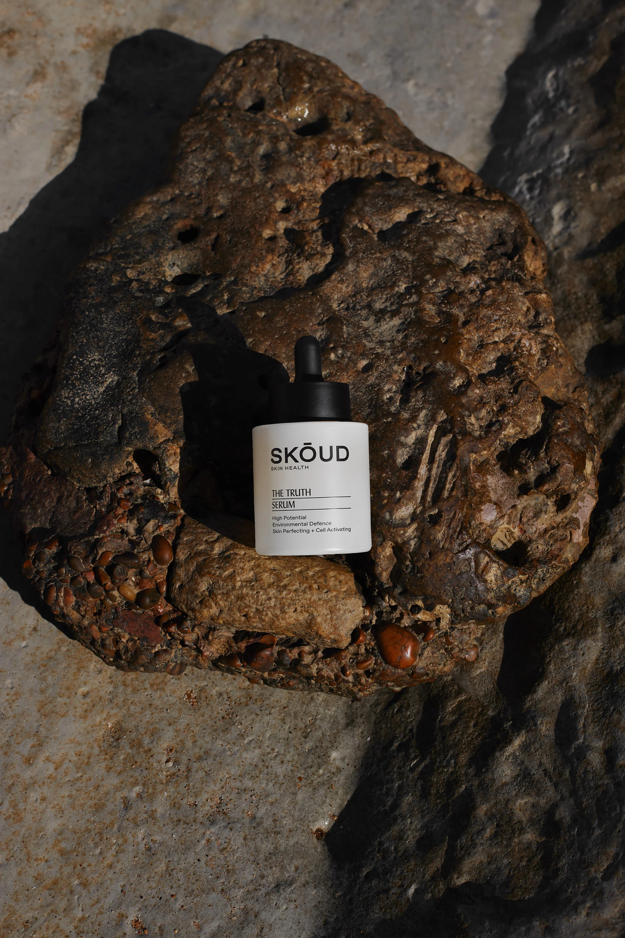 Cosmetic serum bottle, brand name Skoud, sits on a weathered brown rock outdoors, product-focused commercial beauty shot for marketing.