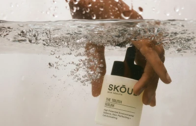 Hand reaching into clear water to hold a Skoud cosmetics skincare serum bottle submerged with air bubbles.