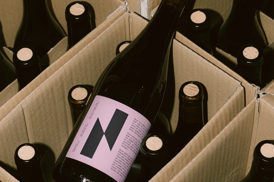 Wine bottles are arranged neatly inside a cardboard box, displaying a pink label with a black geometric design on each bottle.