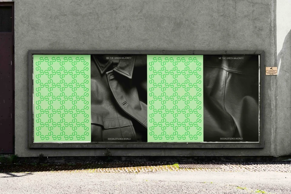 Outdoor billboard with repeating 'S' pattern, jacket close-up, and text 'WE THE GREEN MAJORITY SOCIALSTUDIES.WORLD' on gray concrete wall.