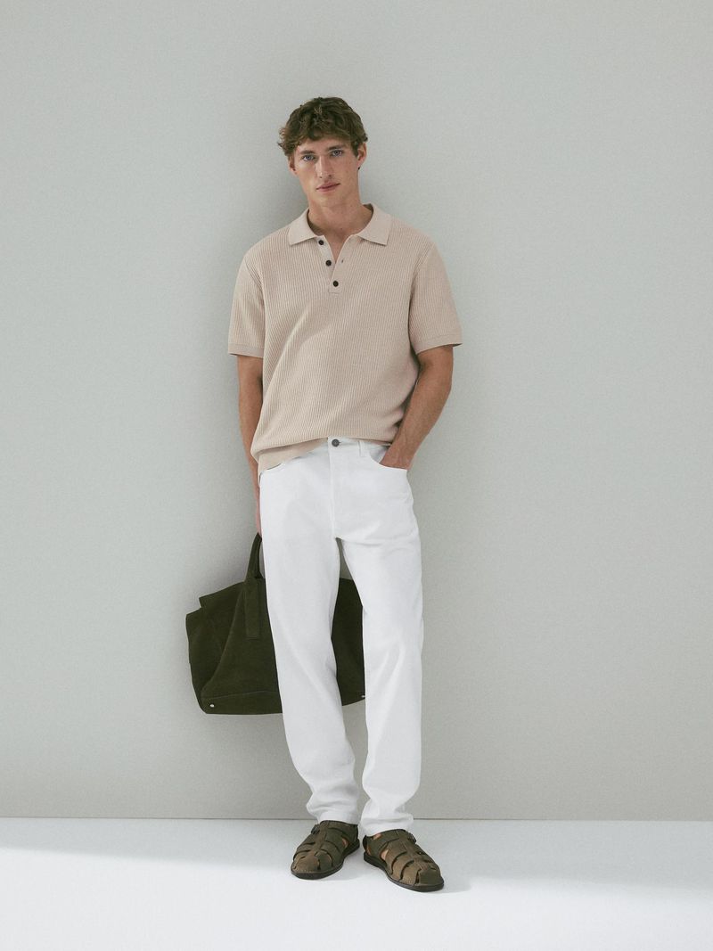Full shot of an adult male wearing a beige polo shirt, white pants, and sandals, holding a green tote bag against a grey wall.