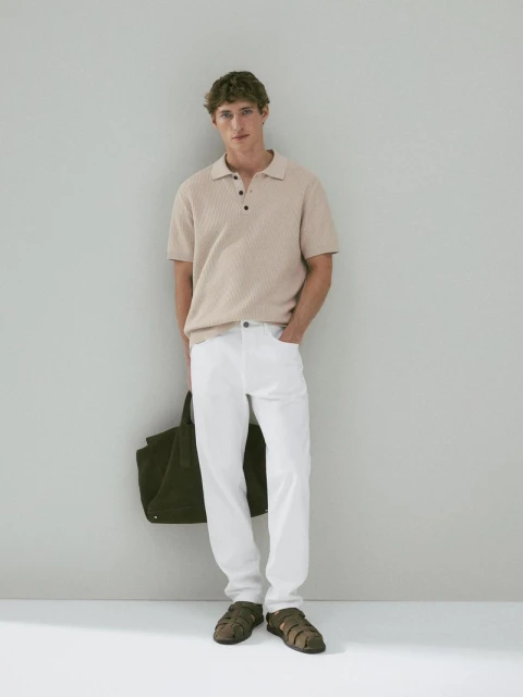 Full shot of an adult male wearing a beige polo shirt, white pants, and sandals, holding a green tote bag against a grey wall.