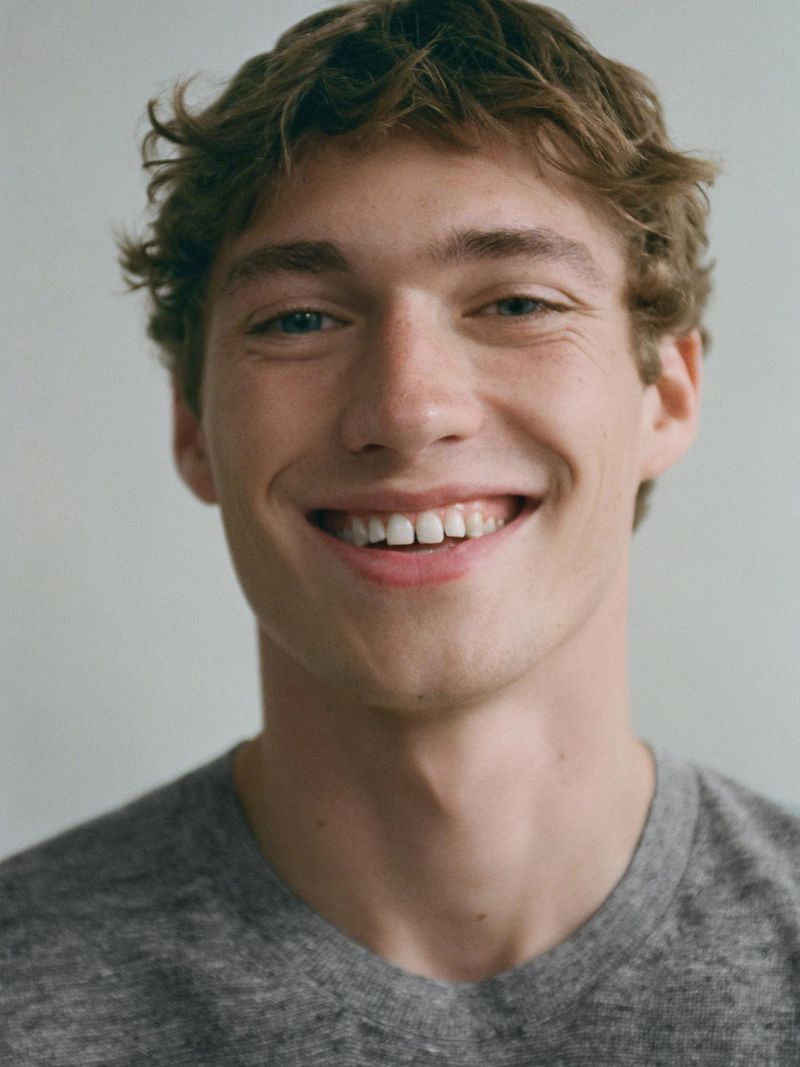 Close-up portrait of a smiling young adult male with curly brown hair, wearing a gray T-shirt, against a neutral background.