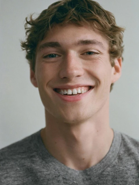 Close-up portrait of a smiling young adult male with curly brown hair, wearing a gray T-shirt, against a neutral background.