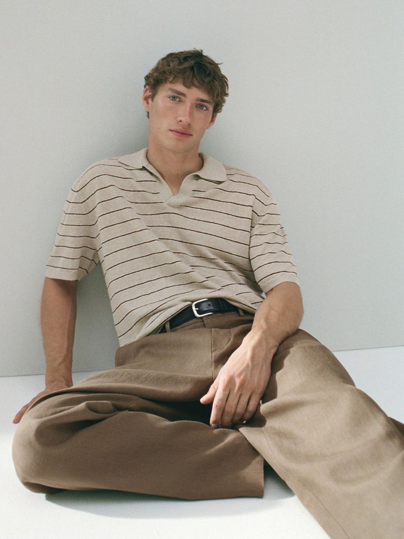 Indoor studio shot of a young adult male model in a striped polo shirt and brown pants sitting against a light wall and floor.