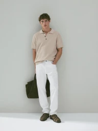 Model wearing a beige knitted polo shirt and white trousers leaning against a seamless grey studio backdrop