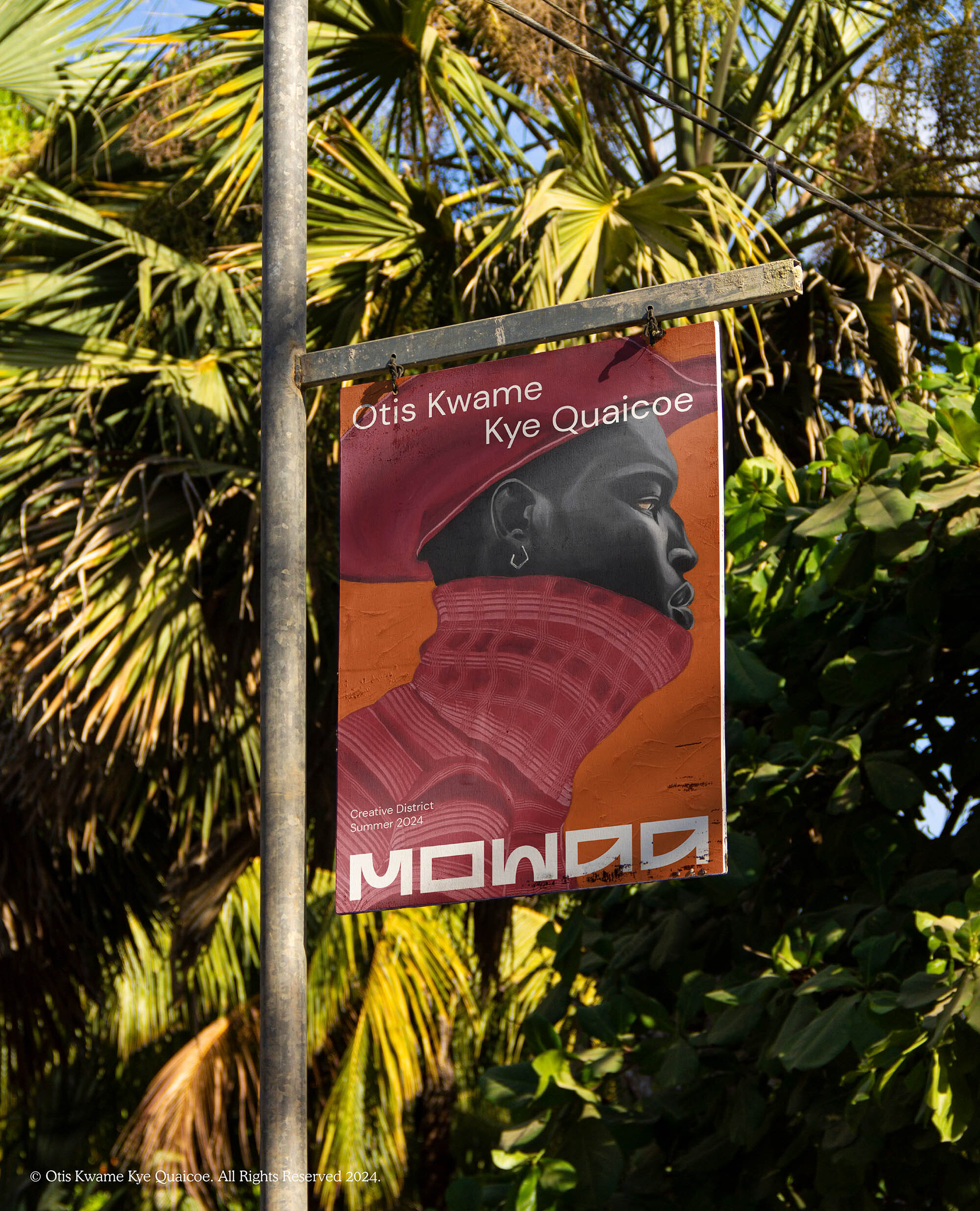 Outdoor sign featuring portrait art of Otis Kwame Kye Quaicoe in red hues, advertising a MOWAA event against a lush green palm backdrop.
