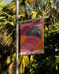 A rectangular sign featuring portrait artwork of a Black man and the MOWAA brand name, mounted on a metal pole amidst green palm leaves.