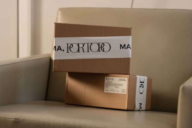Two stacked brown cardboard boxes with 'PORTORO' branding sit on a tan chair, conveying a sense of curated product delivery.