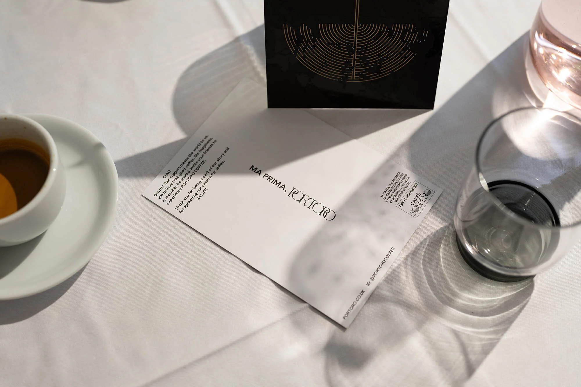 A photo showing a cup of espresso, a small branded card, a coaster, and a glass on a white tablecloth, bathed in natural light.