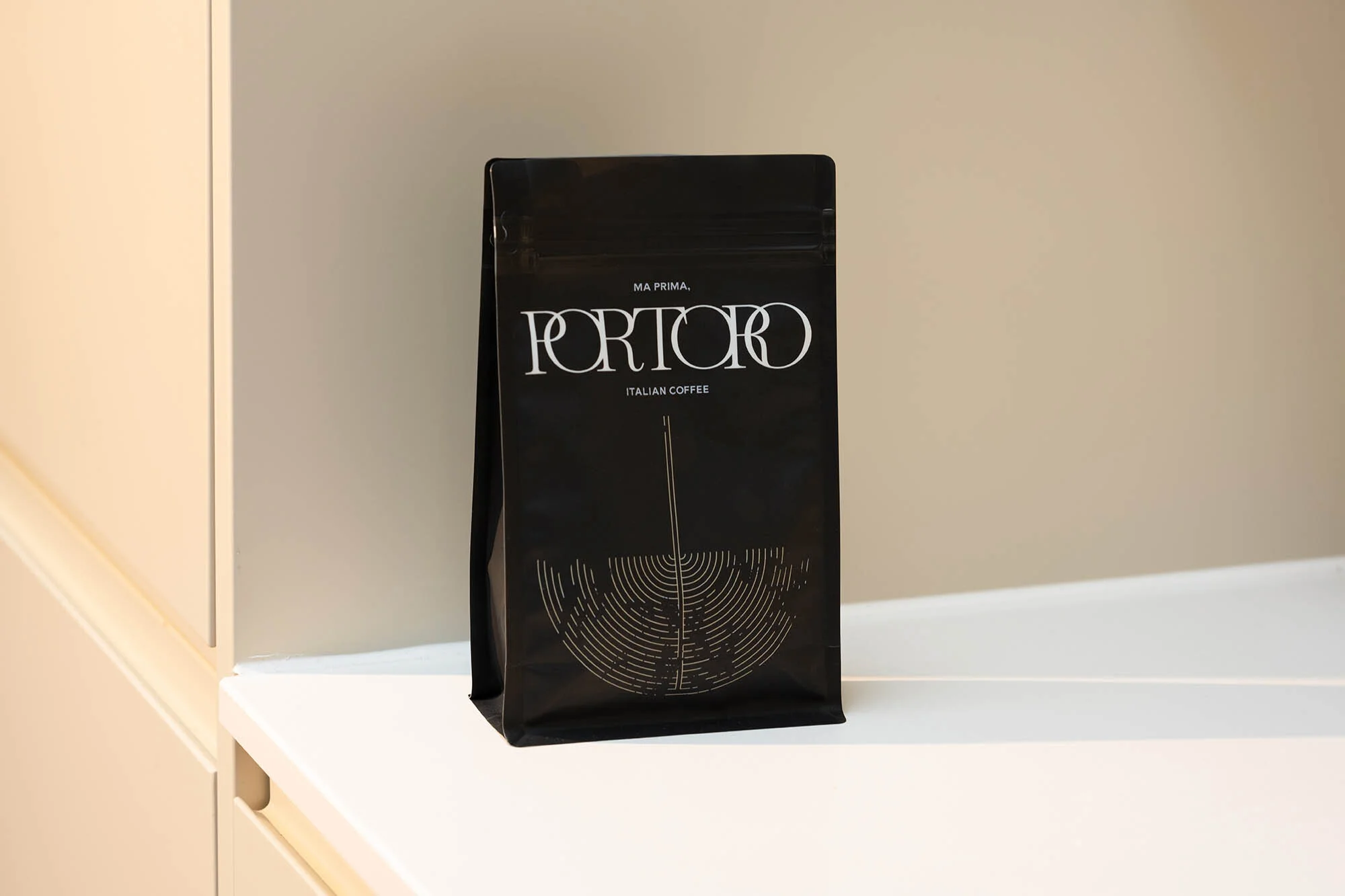 Black coffee bag on a white shelf with 'Portoro Italian Coffee' branding, against a neutral beige wall. Clean product shot.
