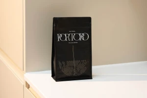 A black coffee bag with white and gold typography resting on a white countertop against a neutral wall.
