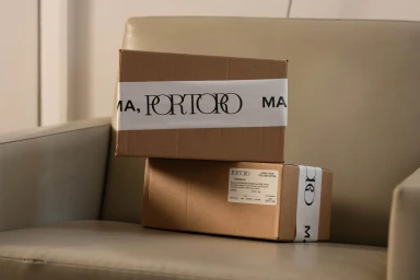 Two stacked cardboard shipping boxes with white branded tape reading Portoro resting on a leather armchair.