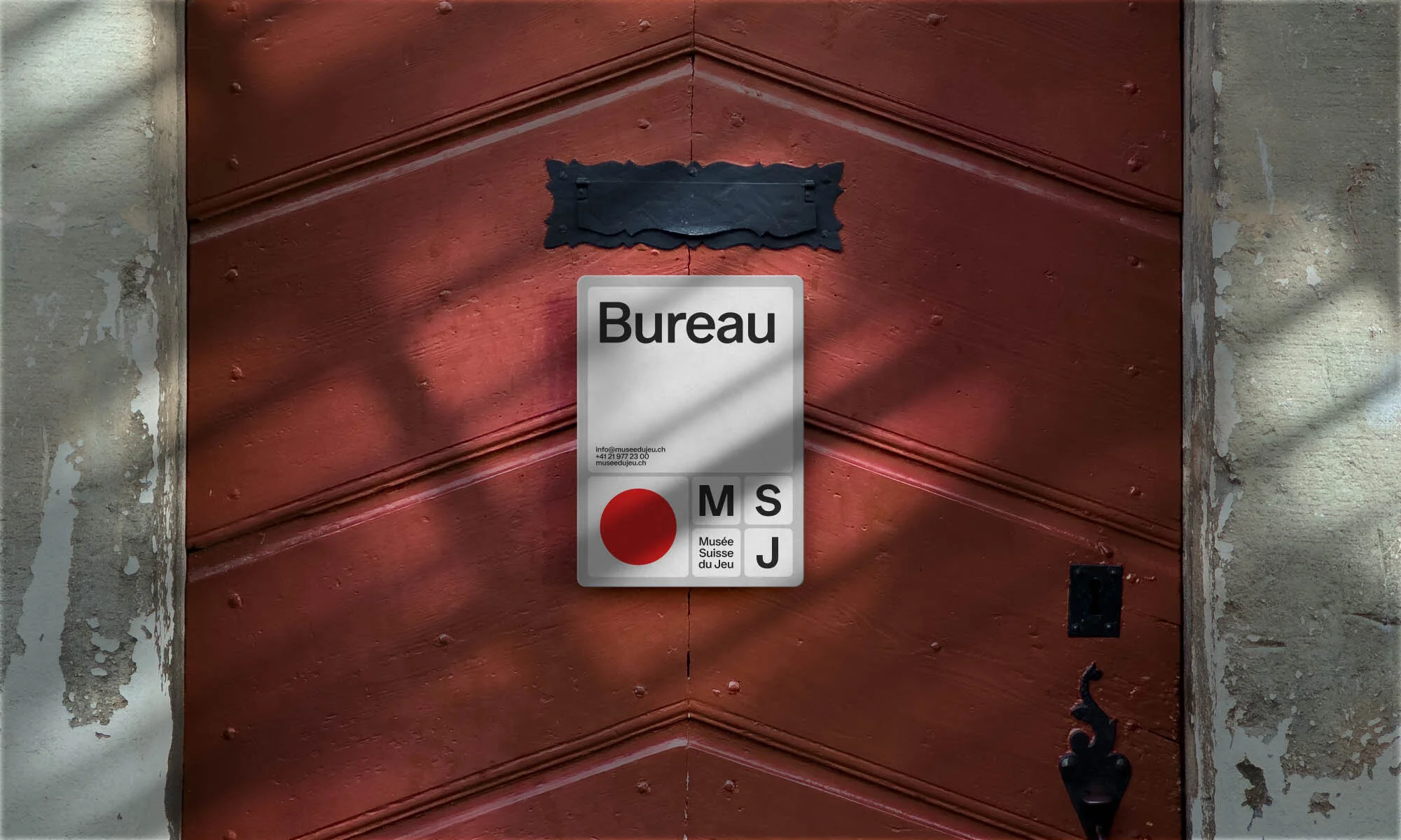 A textured red wooden door displays a square sign for Bureau, featuring a red dot and text for Musée Suisse du Jeu, alongside gray stone.
