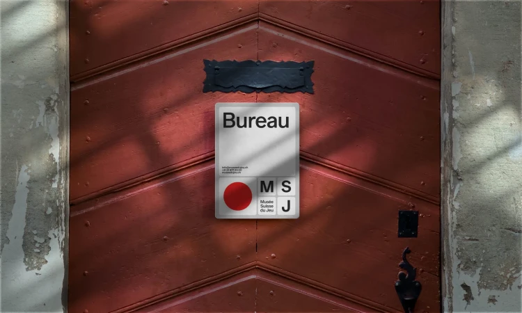 A textured red wooden door displays a square sign for Bureau, featuring a red dot and text for Musée Suisse du Jeu, alongside gray stone.