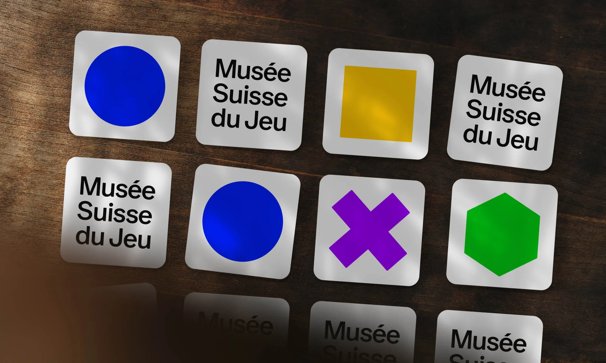 Close-up of game tiles on a dark brown wooden surface, featuring geometric shapes and text 'Musée Suisse du Jeu' repeated.