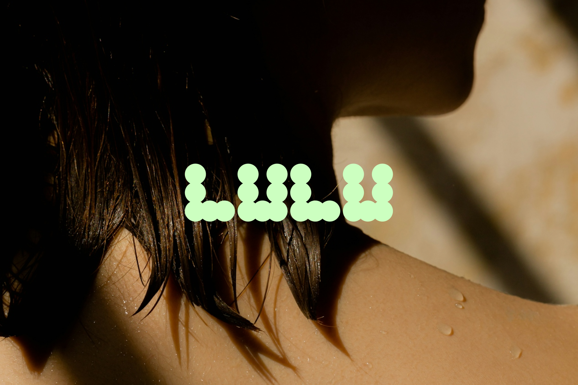 Close-up of a woman's neck and shoulders with wet hair and a light green 'LULU' logo laid across the body in a studio setting.
