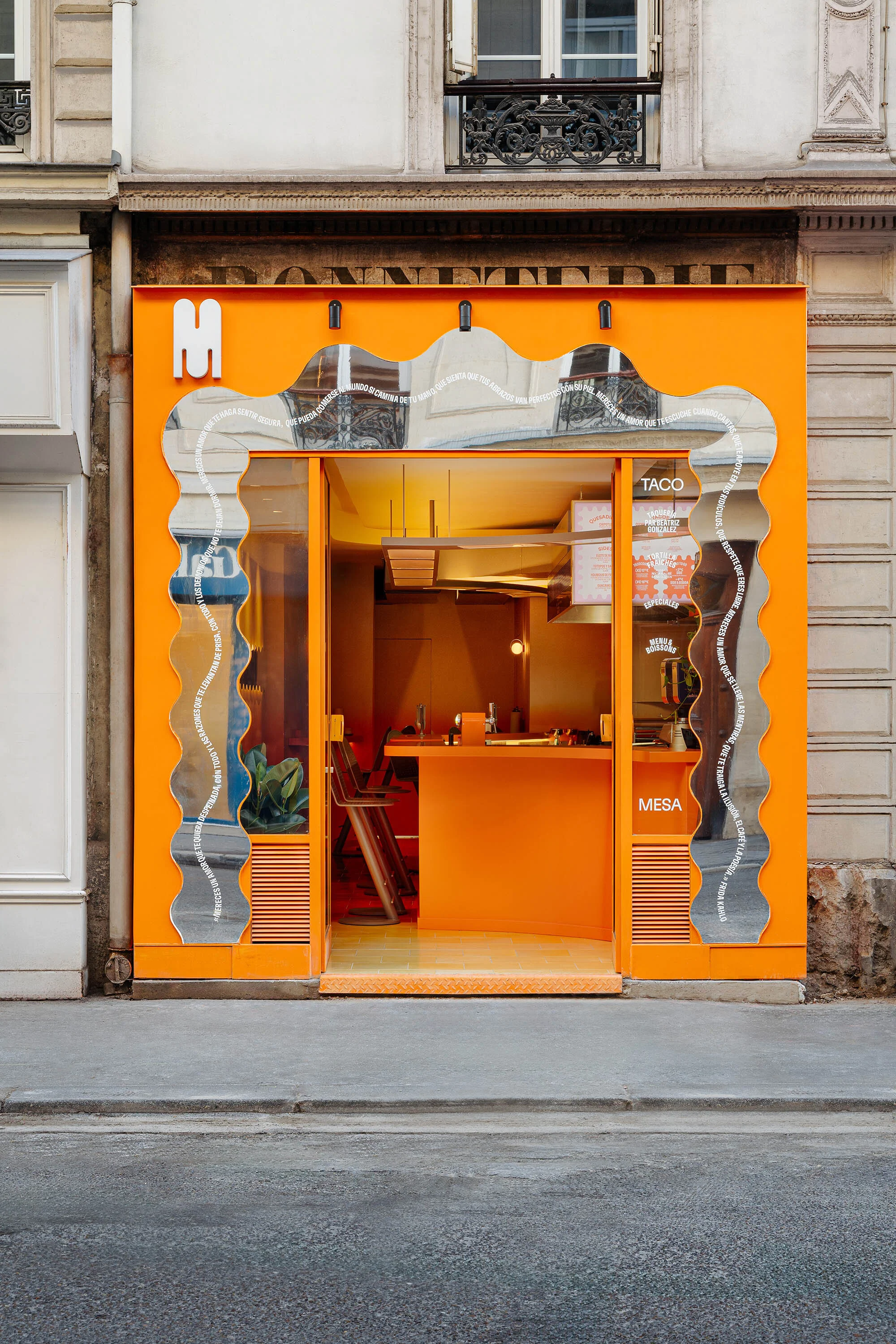 Bright orange exterior of a 'Mesa' taco restaurant in a city setting, featuring a wavy mirror facade and urban street view.