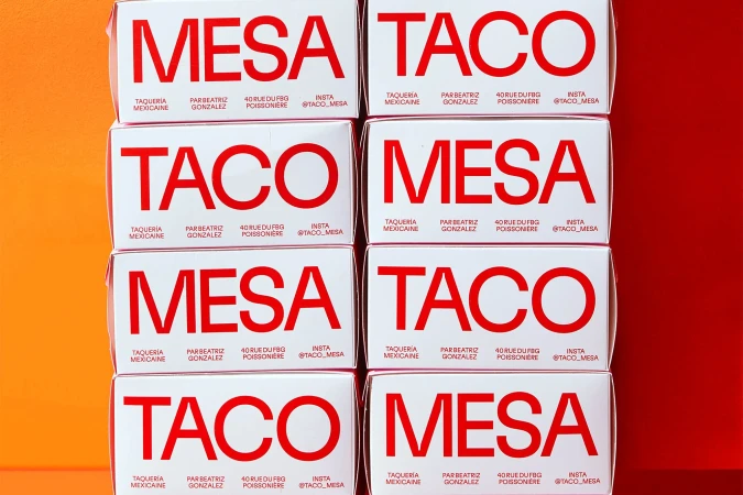 Stacks of white taco boxes with 'Mesa Taco' printed in red, neatly arranged against a contrasting orange and red background.