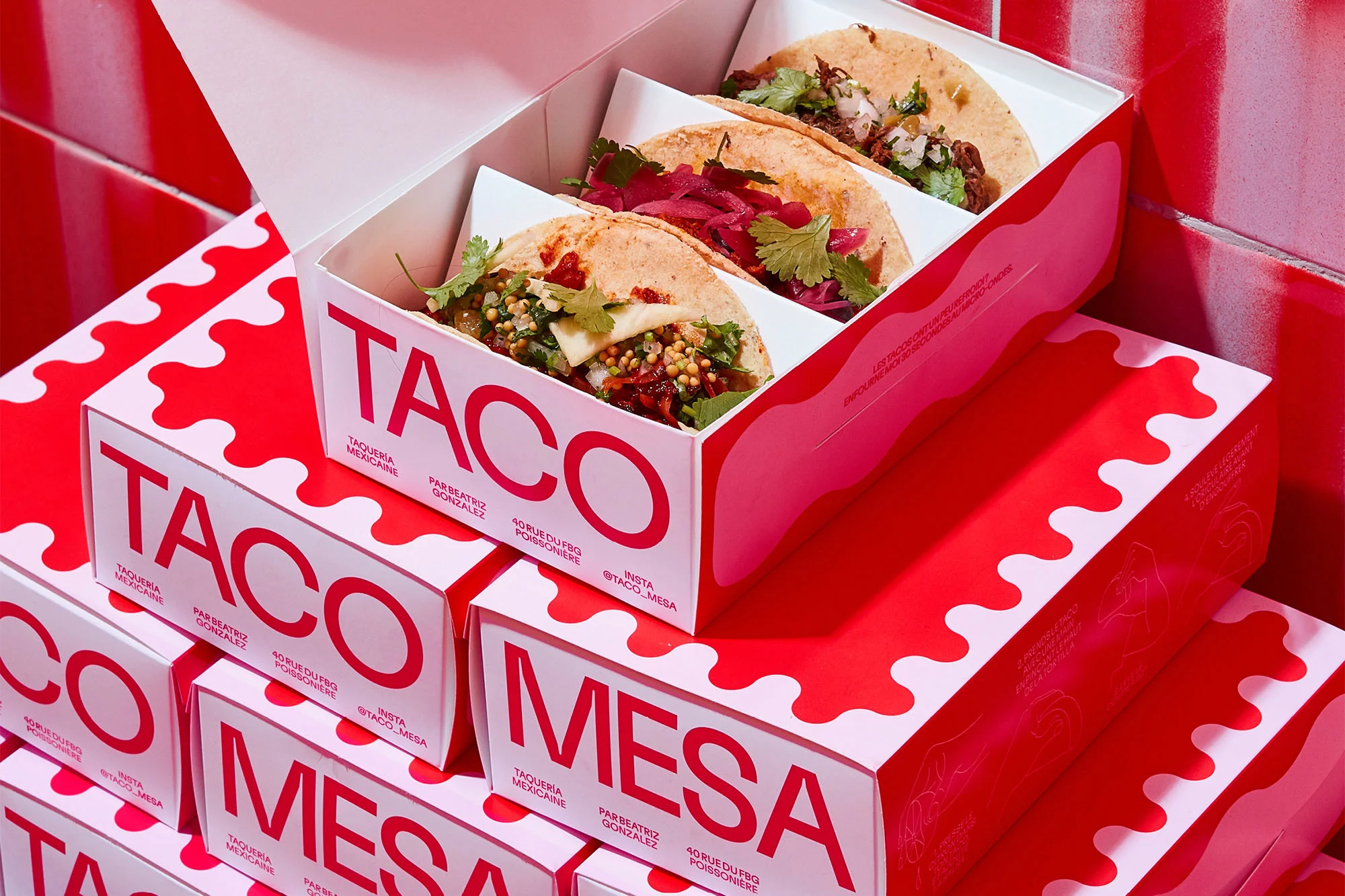 Overhead shot of three gourmet tacos with fresh toppings in an open red and white branded takeout box, stacked with more takeaway boxes.
