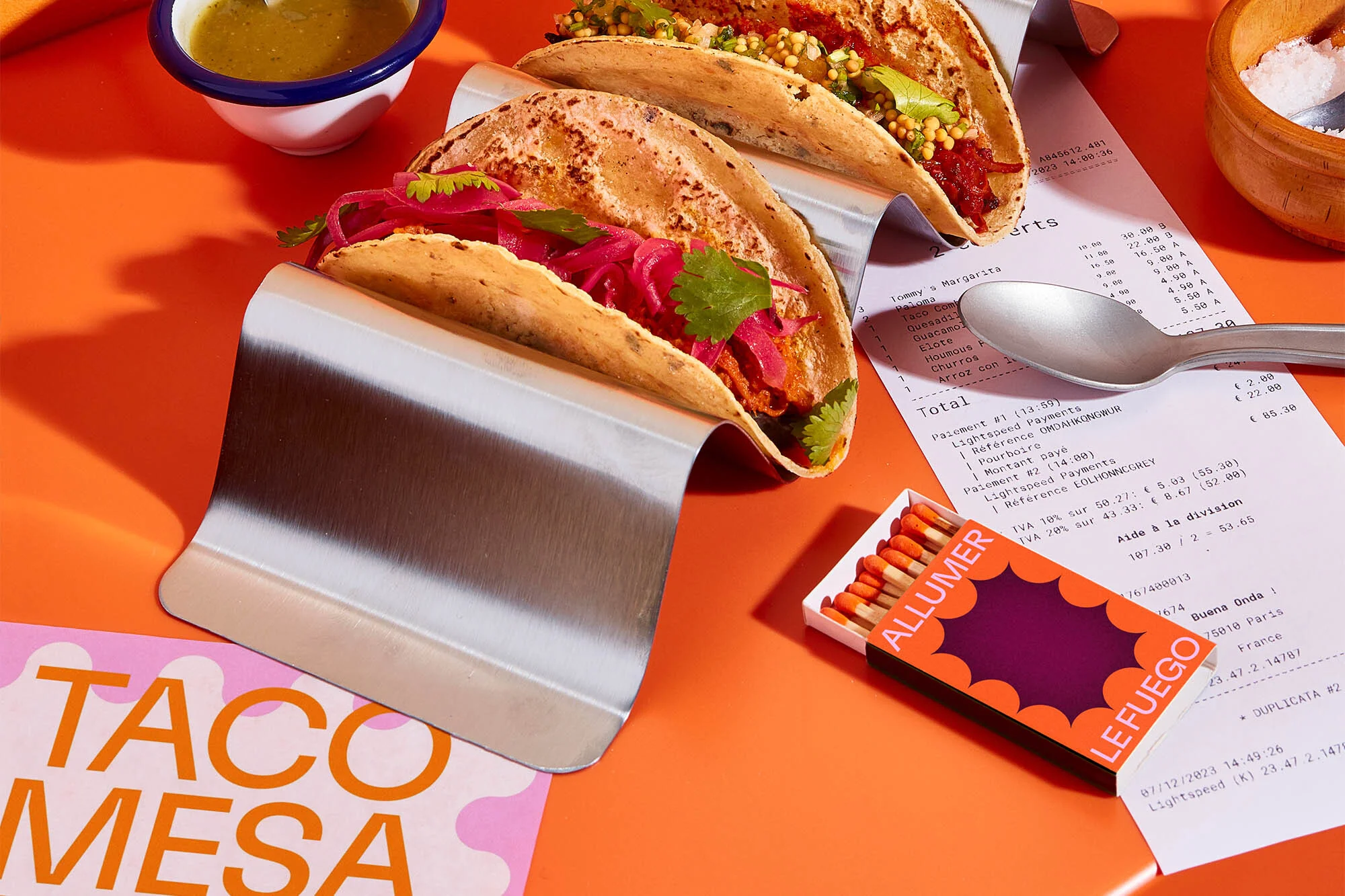 Three tacos in metal holders, side-dish bowl, receipt, matches, salt, and paper card arranged on an orange surface in a studio setting.