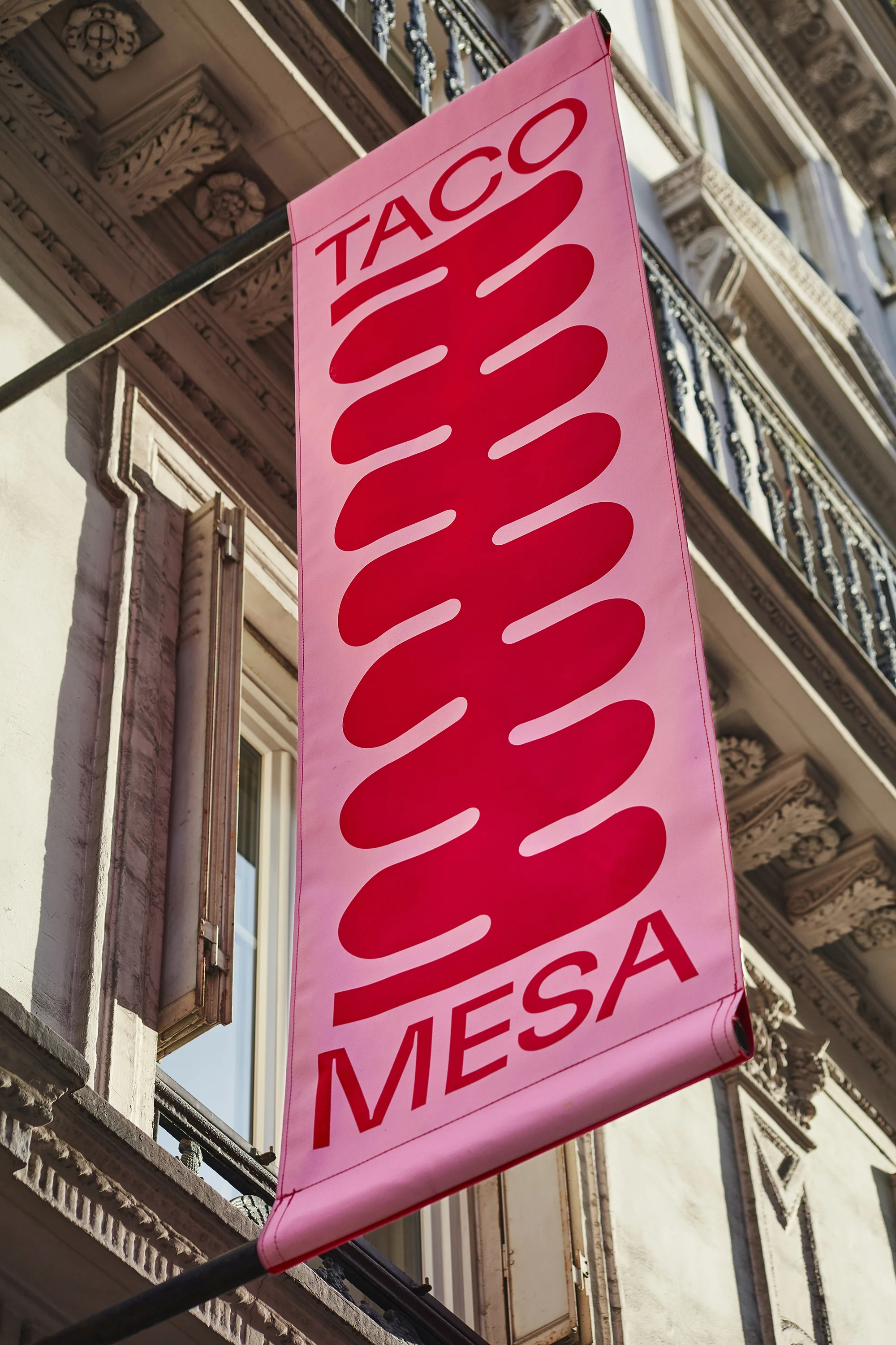 Hanging banner sign for Taco Mesa with abstract, red taco design on a building exterior against a bright, neutral city background.