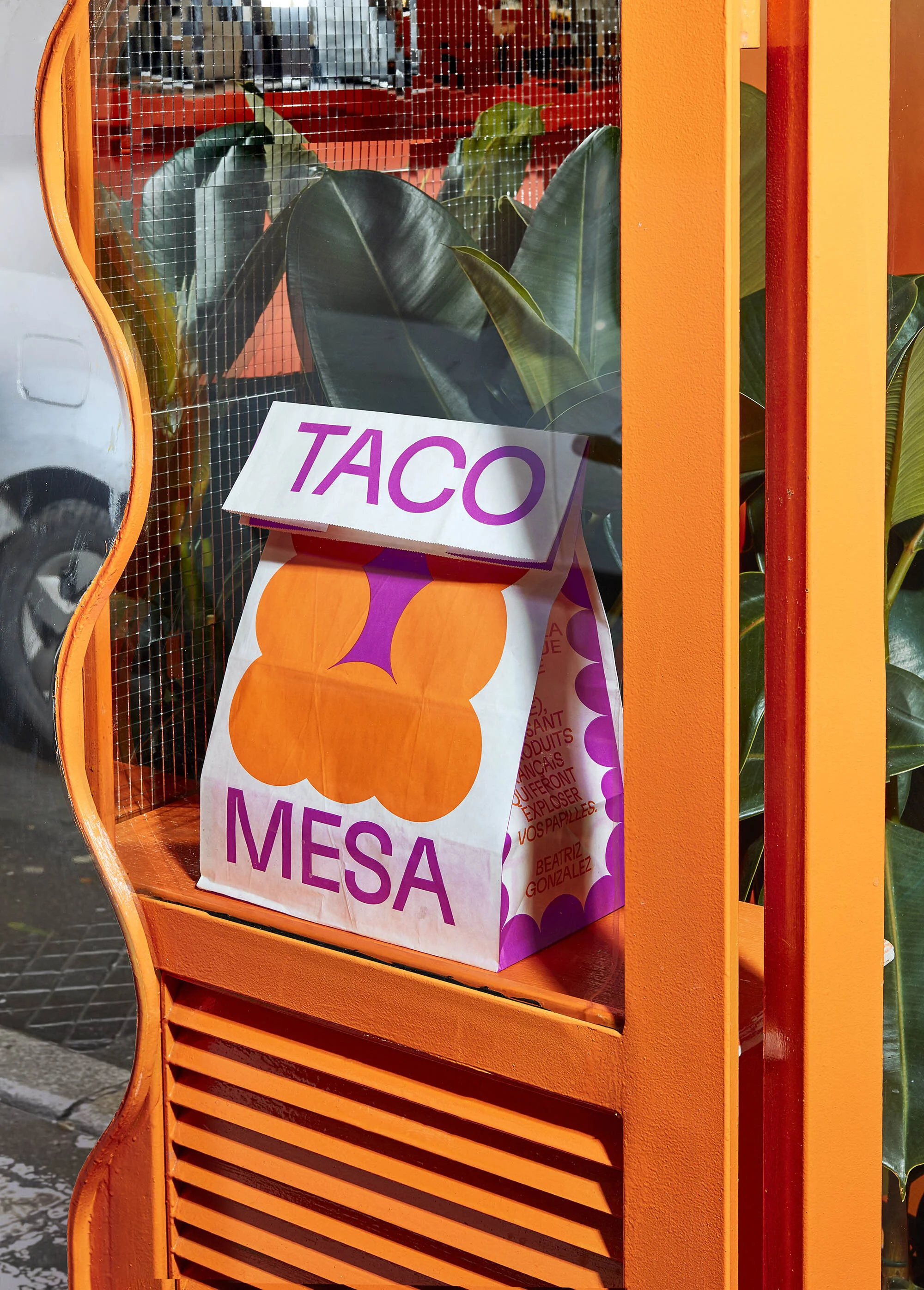 A paper take-out bag with graphic patterns and the word 'TACO' is on display in an orange metal frame, with plant leaves behind.