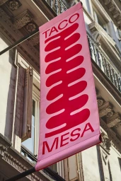 Pink vertical banner flag with red Taco Mesa typography hanging outside a traditional architectural building.