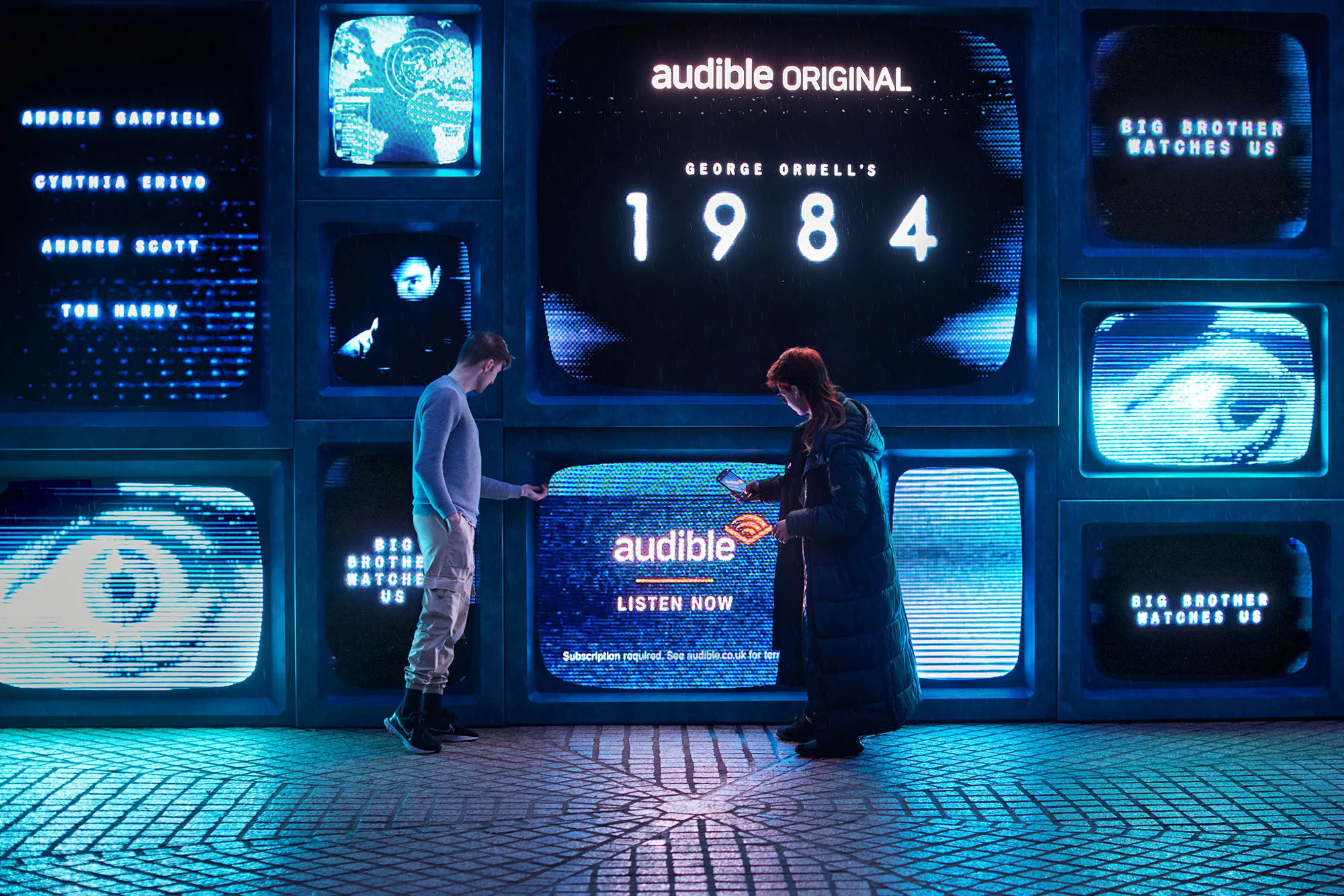 Two people interact with an outdoor Audible advertisement of retro television screens showing George Orwell's 1984 with neon lighting.