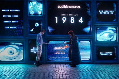 Two people standing before a wall of glowing blue CRT television screens displaying text for Audible Original 1984.