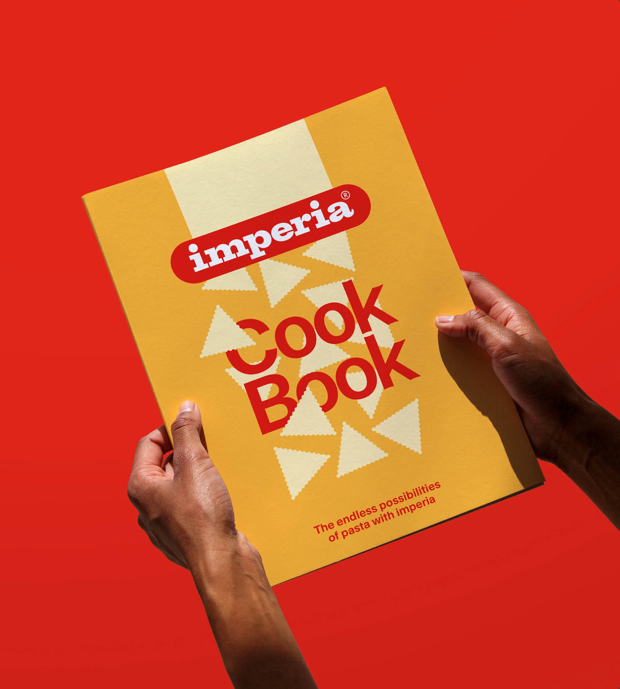Cookbook with Imperia logo held by hands on a red background, featuring a yellow cover with pasta shapes design and brand name.
