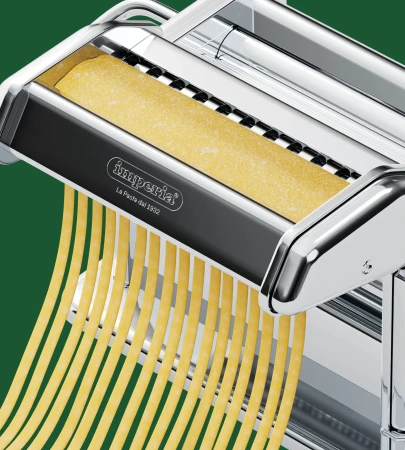 Close-up of a silver Imperia pasta machine creating fresh spaghetti strands on a green backdrop, showcasing kitchen culinary equipment.