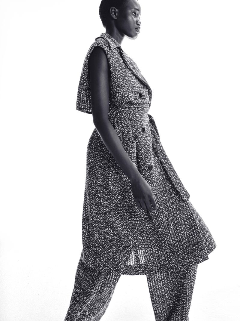 Black and white fashion portrait of a Black female model wearing a tweed-like sleeveless dress with a belt and matching pants in a studio.