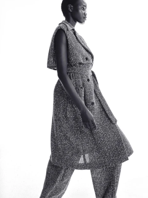 Black and white fashion portrait of a Black female model wearing a tweed-like sleeveless dress with a belt and matching pants in a studio.