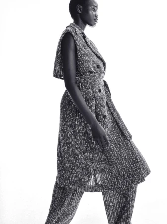 Black and white fashion portrait of a Black female model wearing a tweed-like sleeveless dress with a belt and matching pants in a studio.