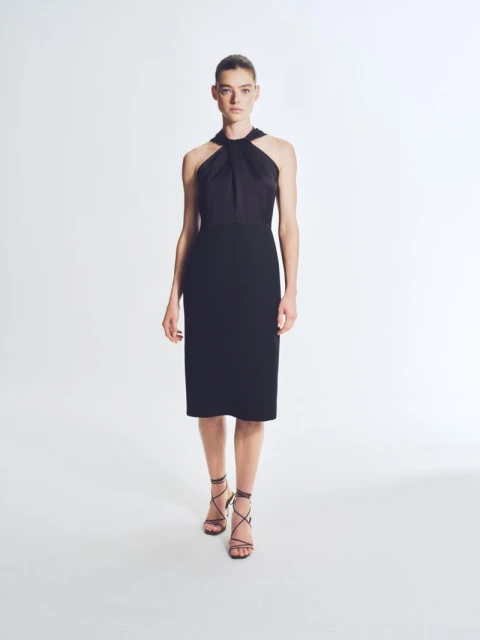 A woman in a black dress and strappy sandals stands in a bright studio with a minimalist backdrop, creating an elegant and modern fashion image.