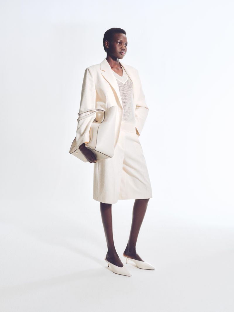 Fashion shot of an adult woman wearing a cream suit with a purse and shoes standing in front of a white background in a photo studio.