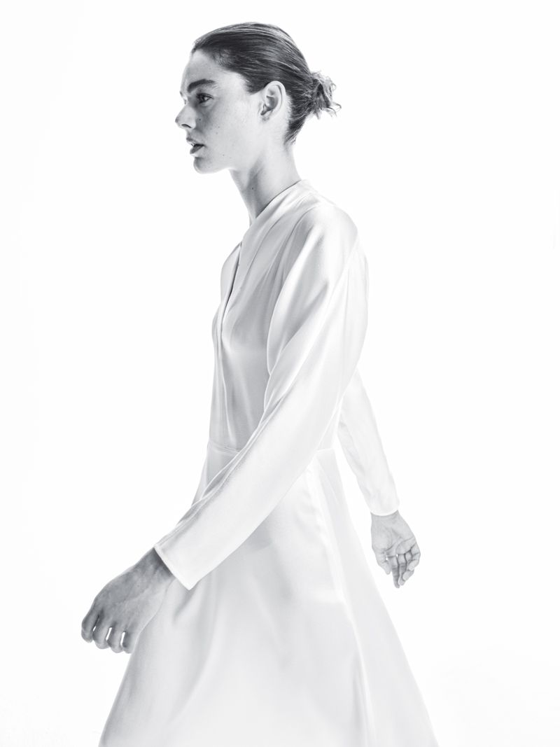 Monochromatic full shot of a slender adult woman walking in a dress with long sleeves against a bright white backdrop.